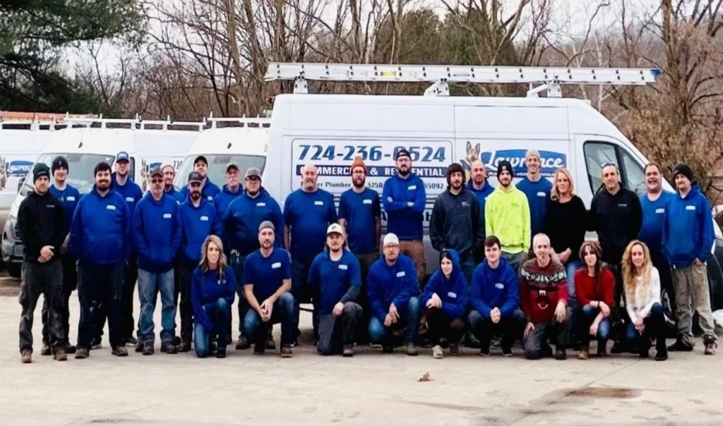 A Group Of People In Blue Work Uniforms Pose In Front Of Two White Service Vans With Company Branding In An Outdoor Setting.